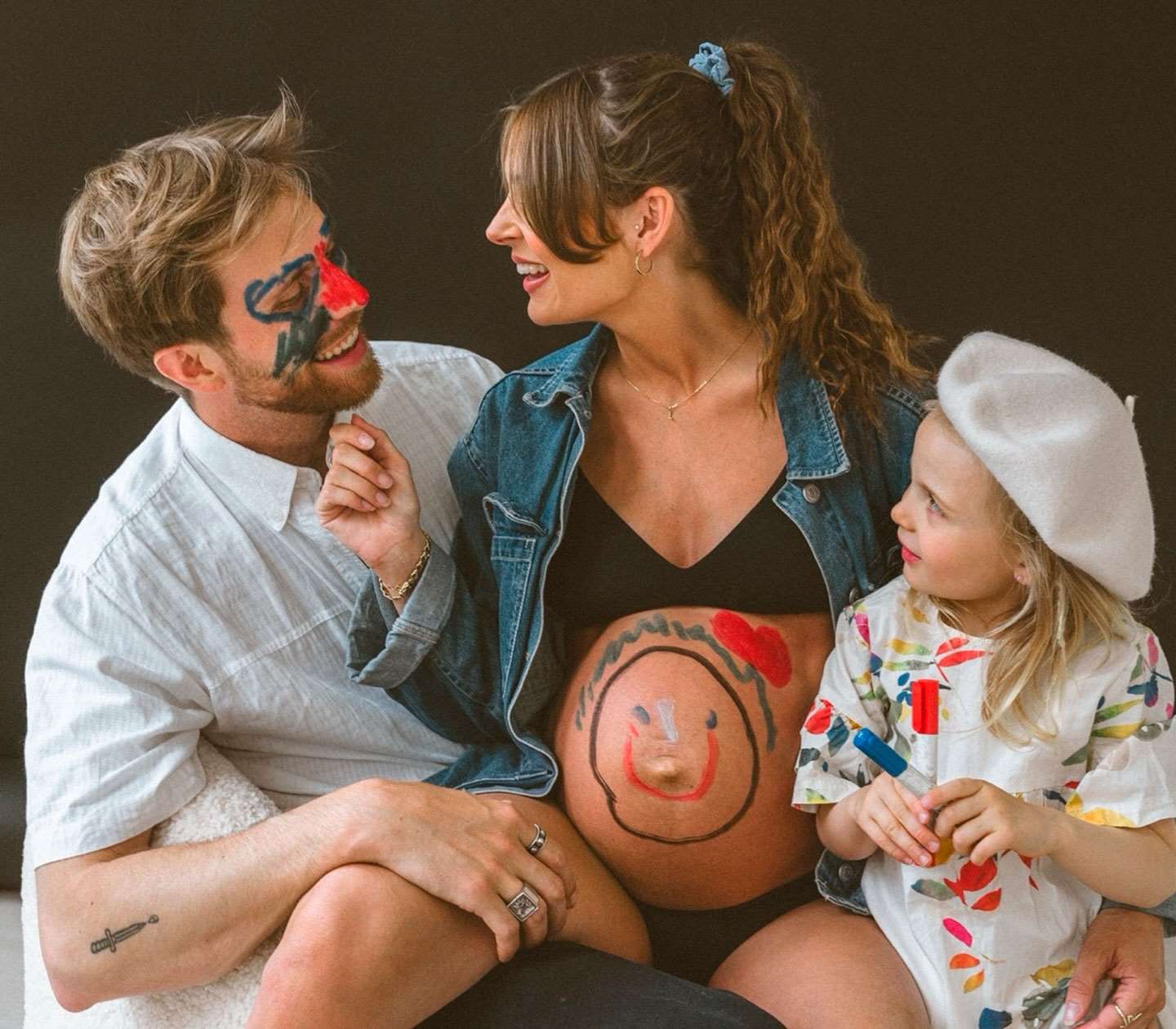 Graviditetsfoto taget af fotograf i Odense. Familie smiler sammen, mens barnet har tegnet på fars ansigt og mors mave.