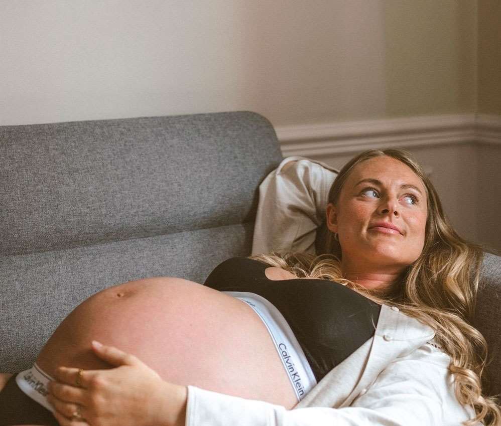 Graviditetsportræt taget af fotograf i Odense. Kvinde hviler på sofaen med hånden på sin mave og et roligt smil.